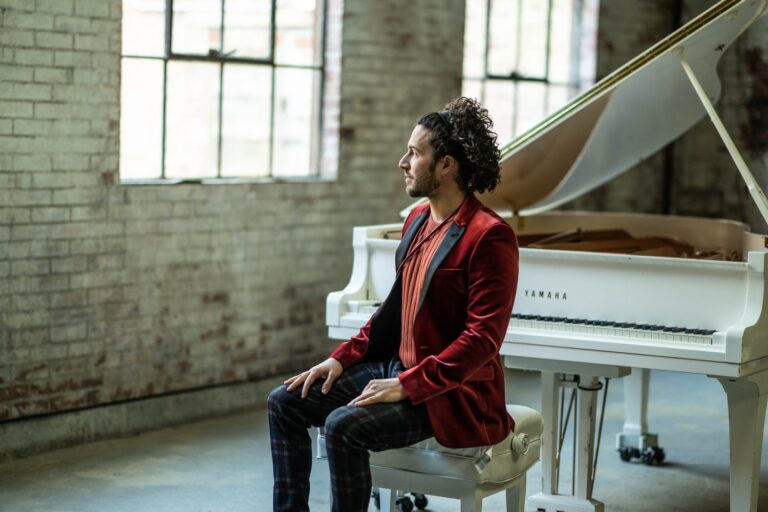 Pianist, Emmet Cohen sitting on a piano bench looking to the left.